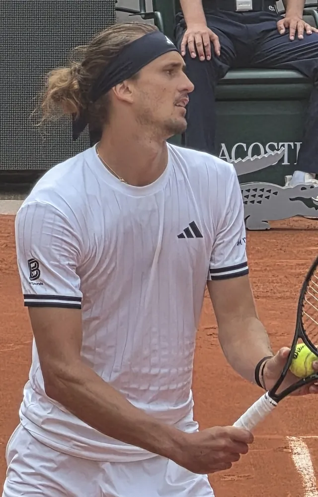Alexander Zverev im weißen Shirt mit Stirnband und Zopf hält Ball und Schläger vor dem Aufschlag, Roland Garros 2026